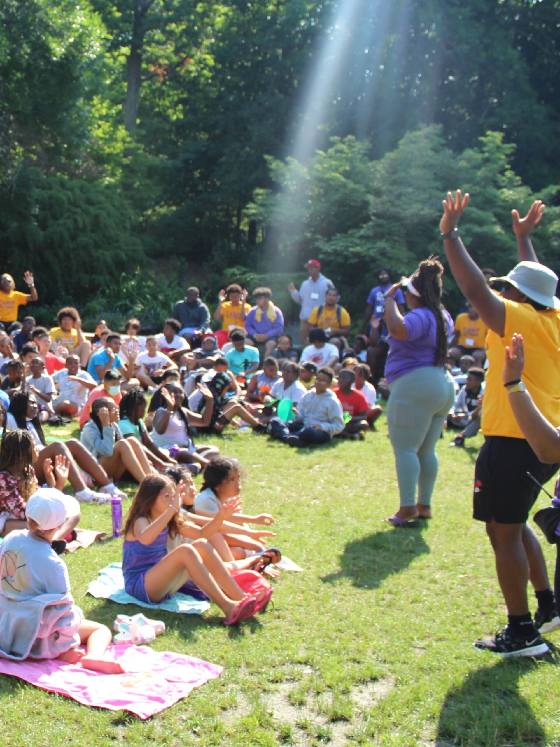 Youth sitting on the ground while adults address them and cheer.